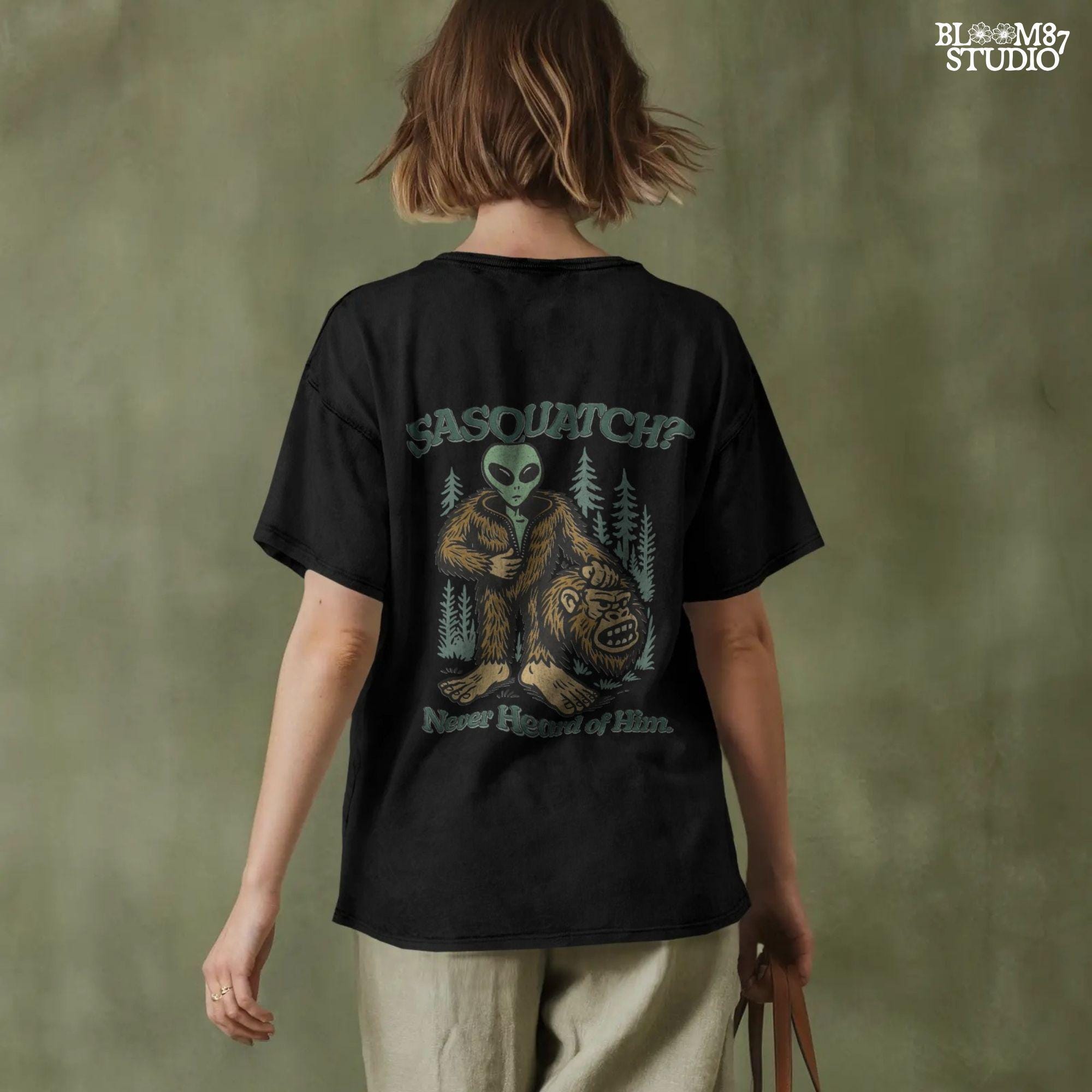 Funny alien unzipping a Bigfoot costume with Sasquatch head in hand, forest trees in background, and text “Sasquatch? Never Heard of Him.”