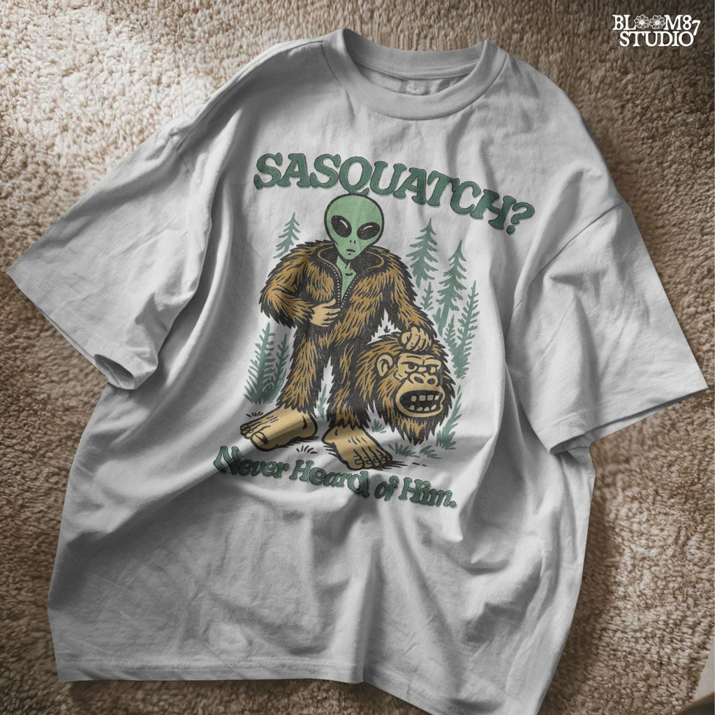 Funny alien unzipping a Bigfoot costume with Sasquatch head in hand, forest trees in background, and text “Sasquatch? Never Heard of Him.”