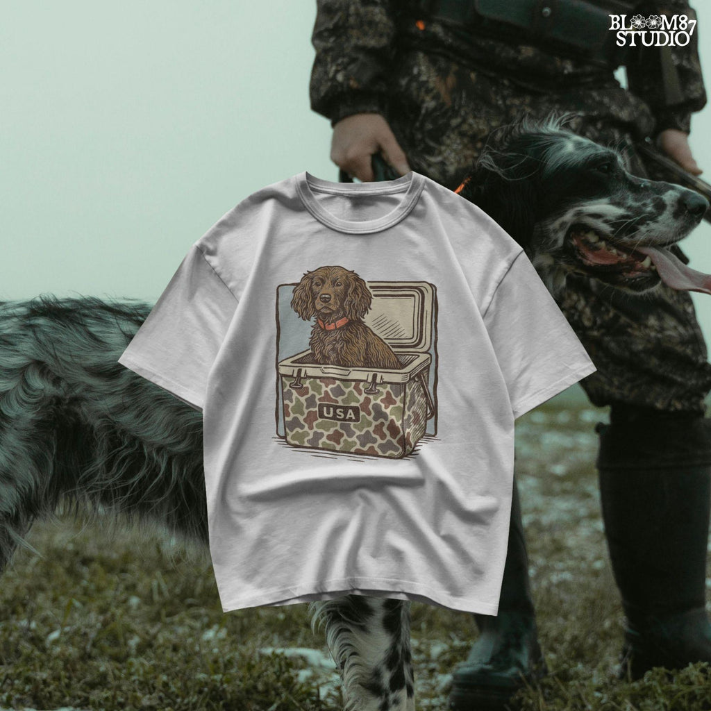 Hunting dog with curly fur sitting inside a camouflage cooler marked “USA” in a retro-style outdoors graphic.