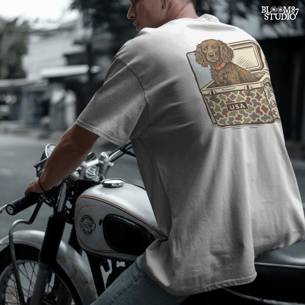 Hunting dog with curly fur sitting inside a camouflage cooler marked “USA” in a retro-style outdoors graphic.