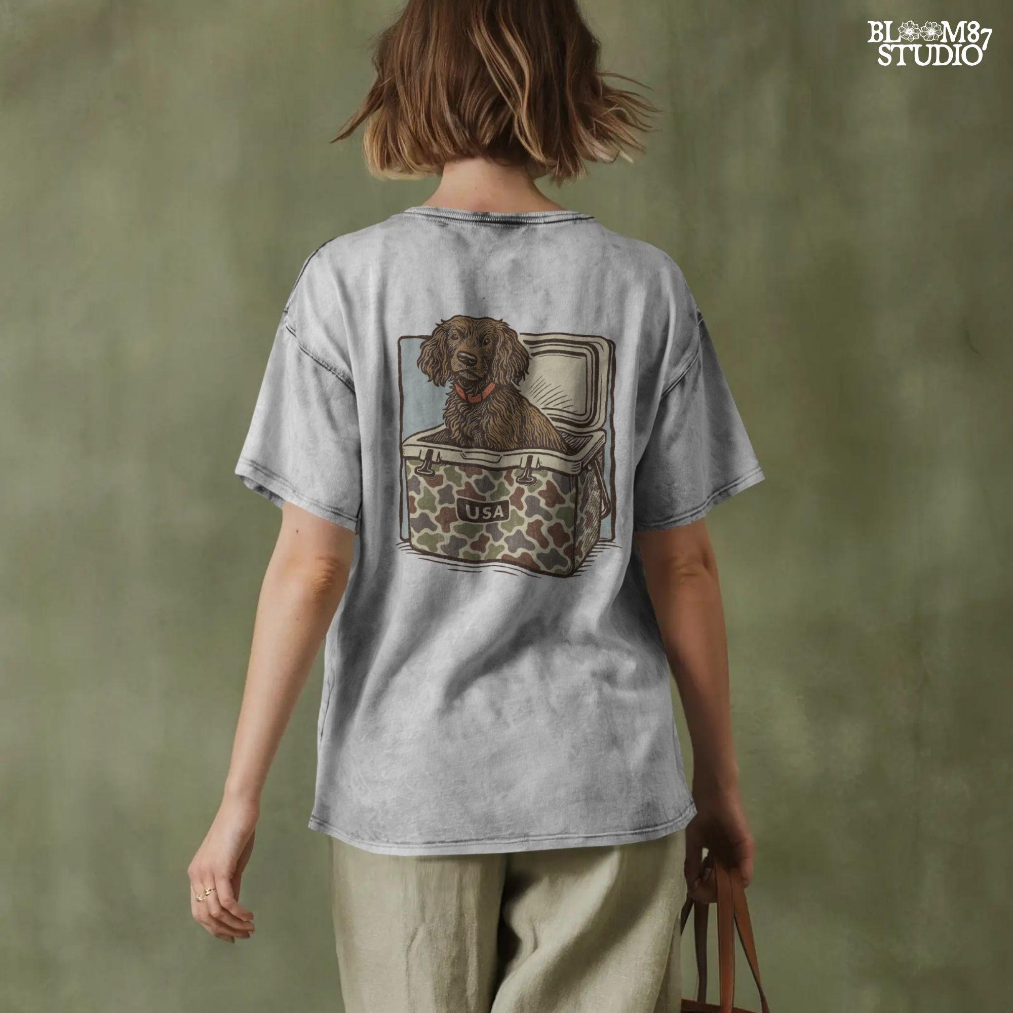 Hunting dog with curly fur sitting inside a camouflage cooler marked “USA” in a retro-style outdoors graphic.