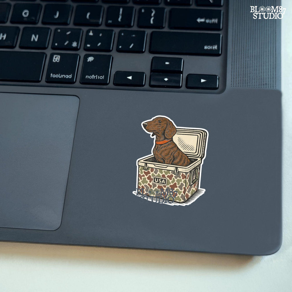 Brown dachshund dog sitting in camouflage cooler labeled “USA” with a red collar, drawn in patriotic cartoon style