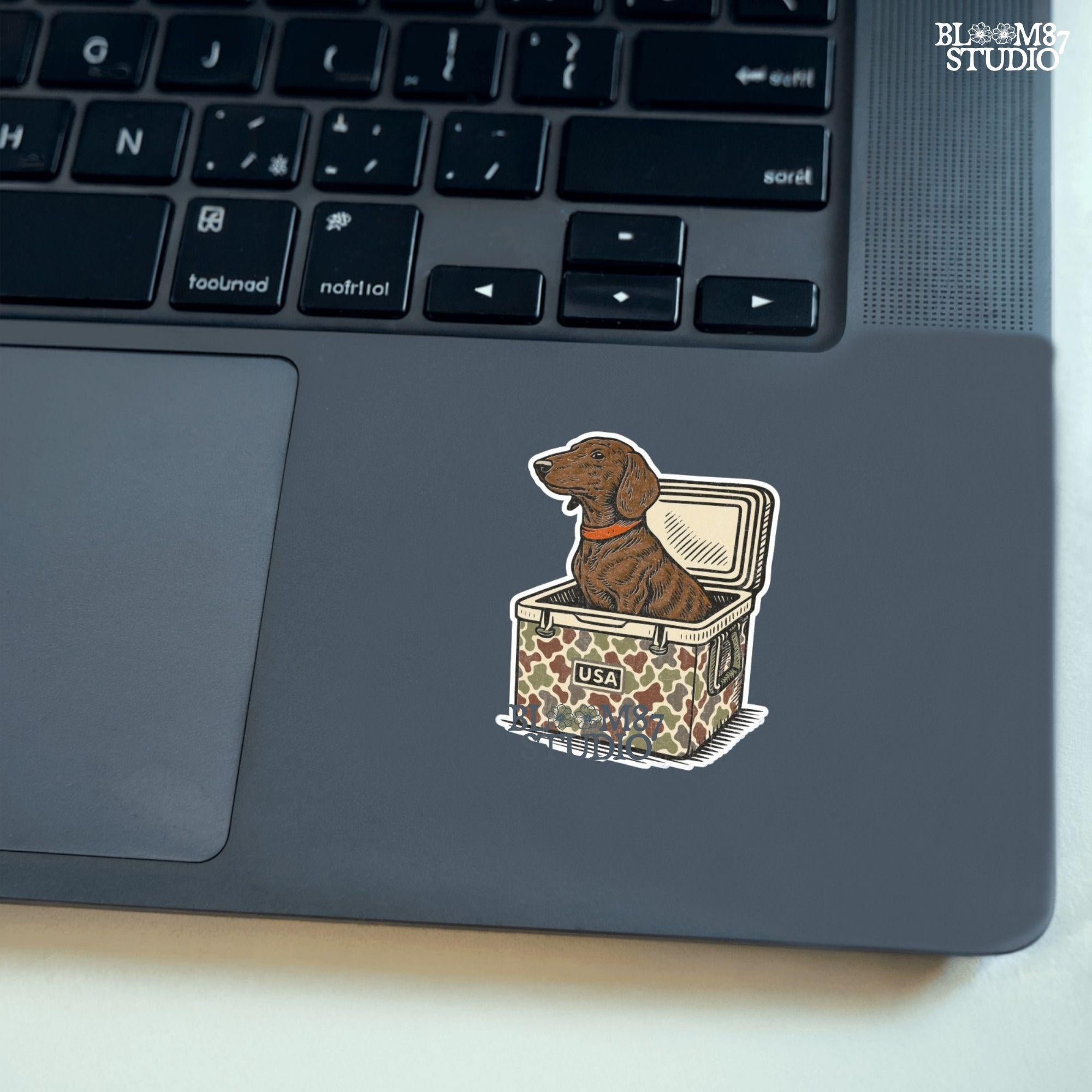 Brown dachshund dog sitting in camouflage cooler labeled “USA” with a red collar, drawn in patriotic cartoon style