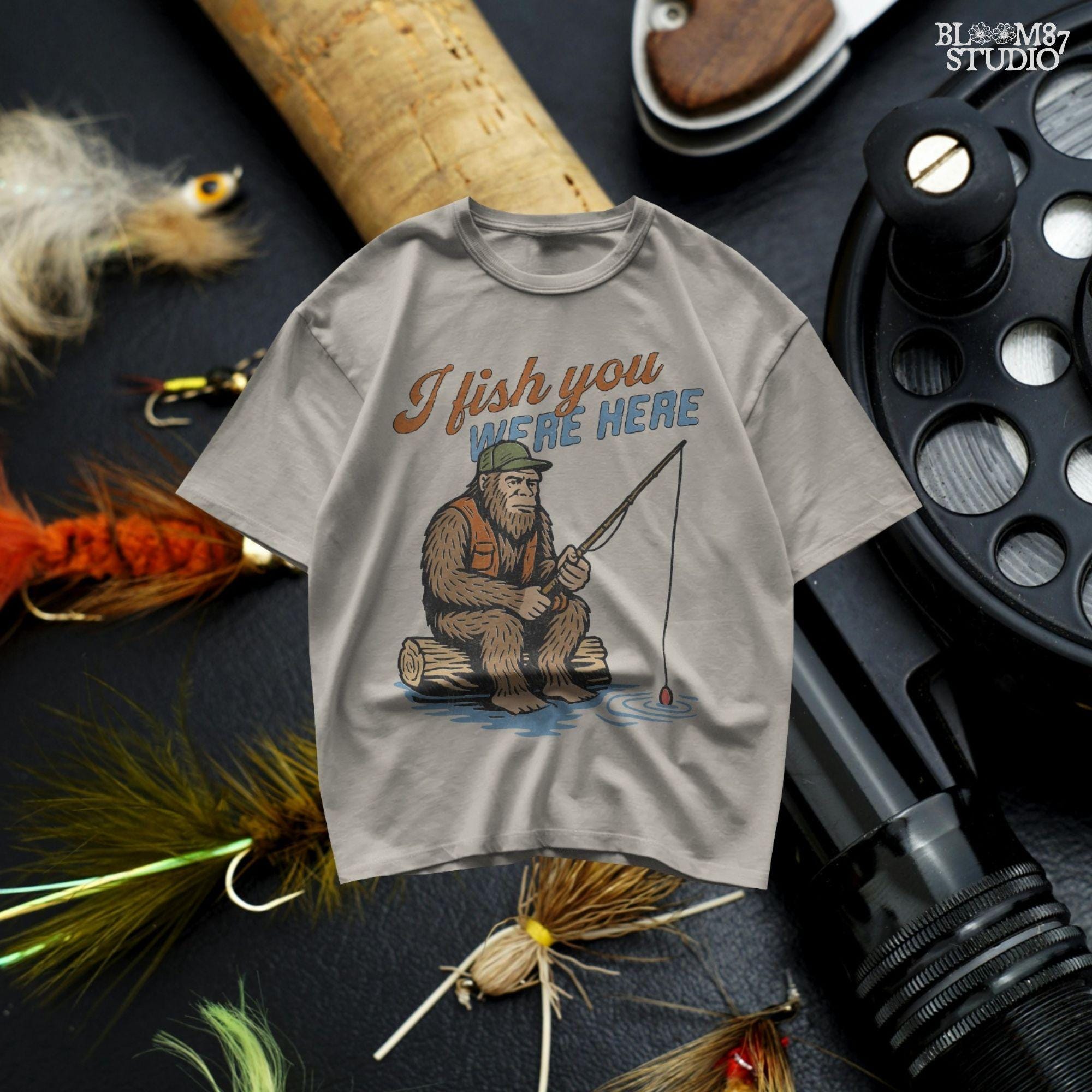 Funny Bigfoot character sitting on log fishing with bobber, wearing orange vest and green cap, with text “I Fish You Were Here”