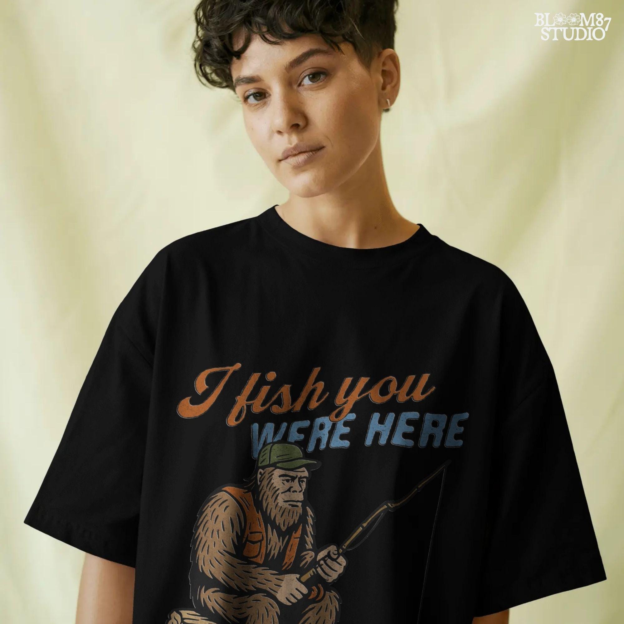 Funny Bigfoot character sitting on log fishing with bobber, wearing orange vest and green cap, with text “I Fish You Were Here”