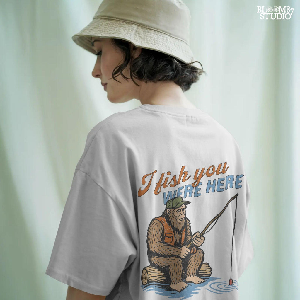 Funny Bigfoot character sitting on log fishing with bobber, wearing orange vest and green cap, with text “I Fish You Were Here”