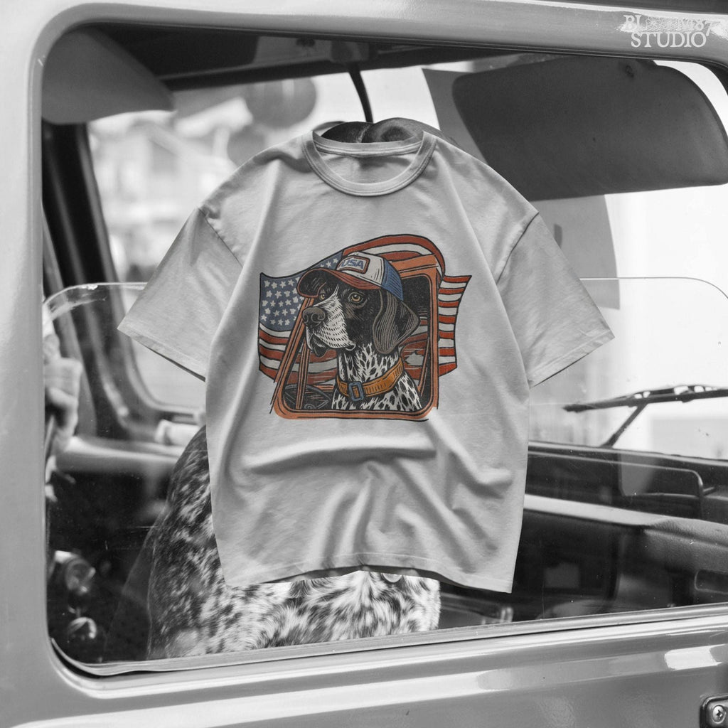 German Shorthaired Pointer dog wearing USA hat looking out of truck window with American flag waving in background