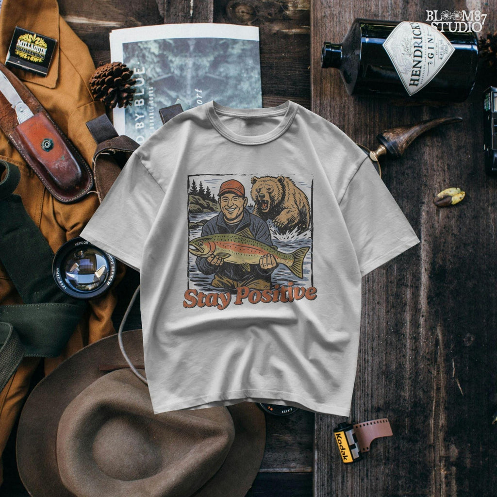 Happy fisherman holding large trout in river while a grizzly bear charges from behind, with text “Stay Positive” in funny outdoor design style