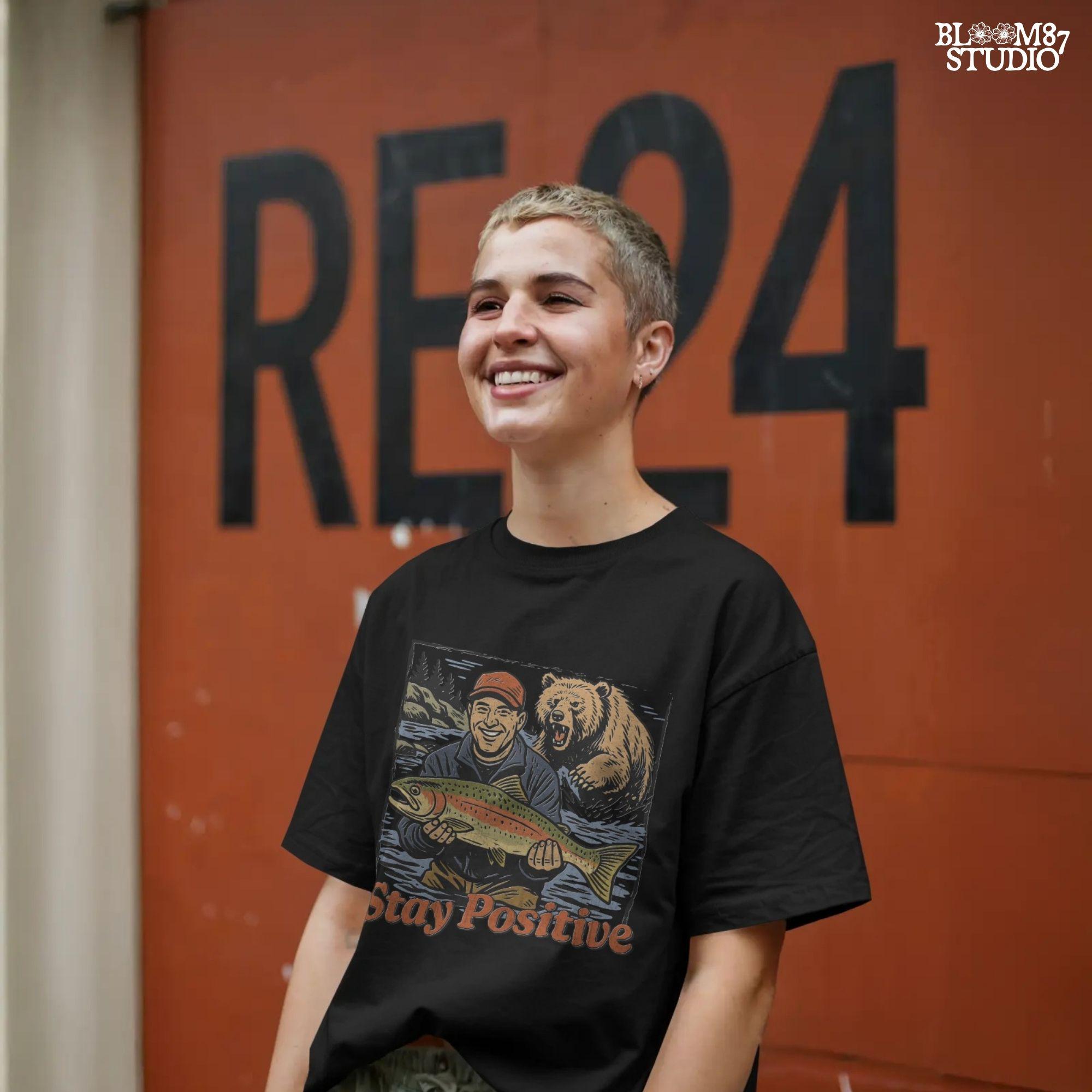 Happy fisherman holding large trout in river while a grizzly bear charges from behind, with text “Stay Positive” in funny outdoor design style