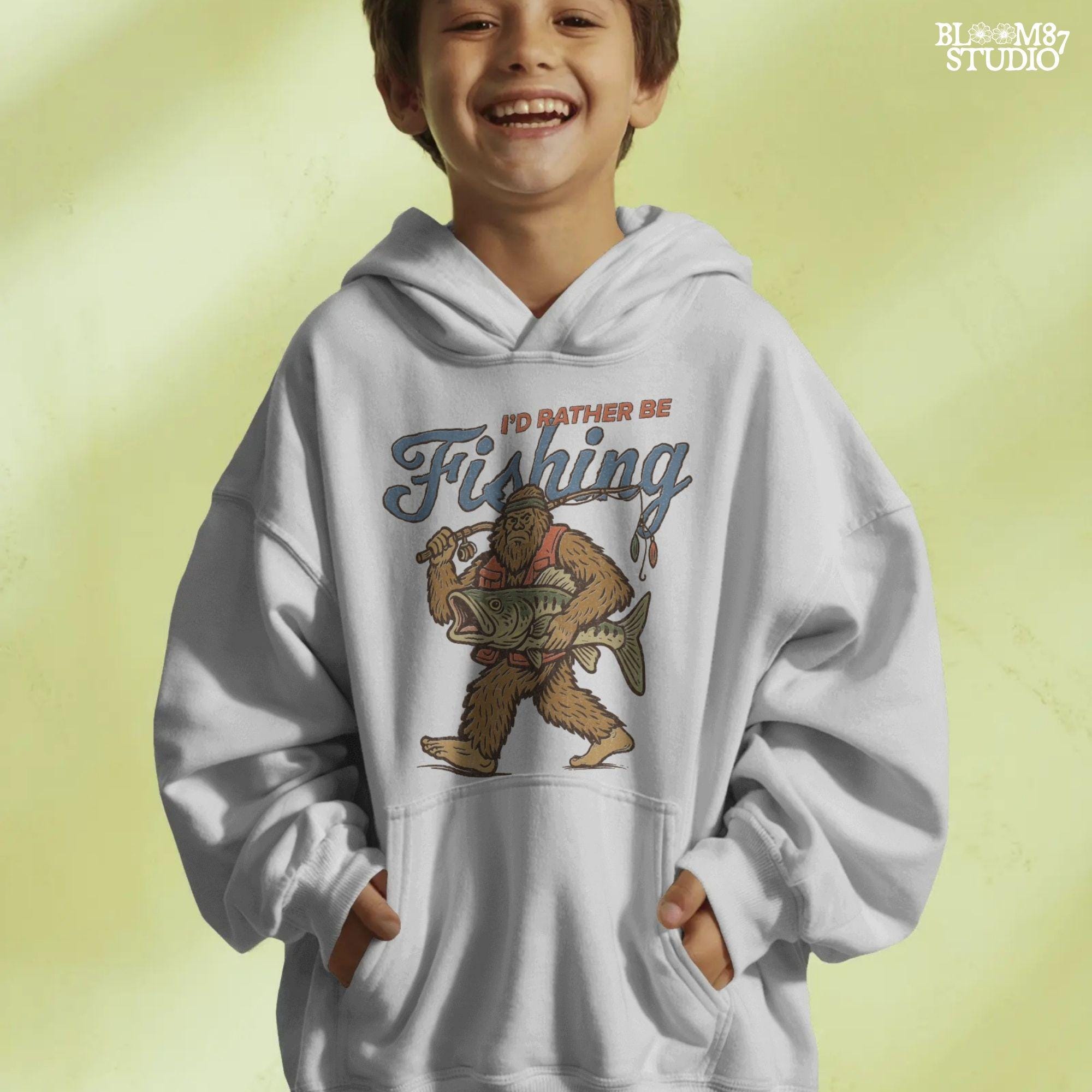 Funny vintage illustration of Bigfoot wearing a fishing vest, holding a giant bass and fishing rod, with text that reads “I’d Rather Be Fishing.”