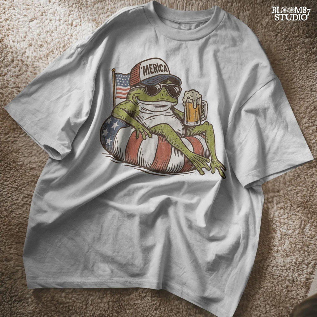 A vintage-style cartoon frog lounging on a patriotic inner tube, wearing a ‘Merica cap and sunglasses, holding a mug of beer with an American flag in the background.