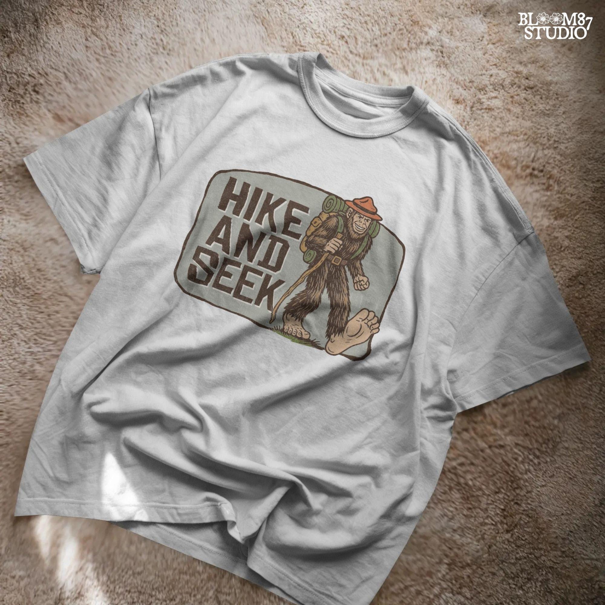 Illustration of a smiling Bigfoot wearing a ranger hat and hiking backpack with the phrase “Hike and Seek,” designed as a PNG for sublimation or print-on-demand products.