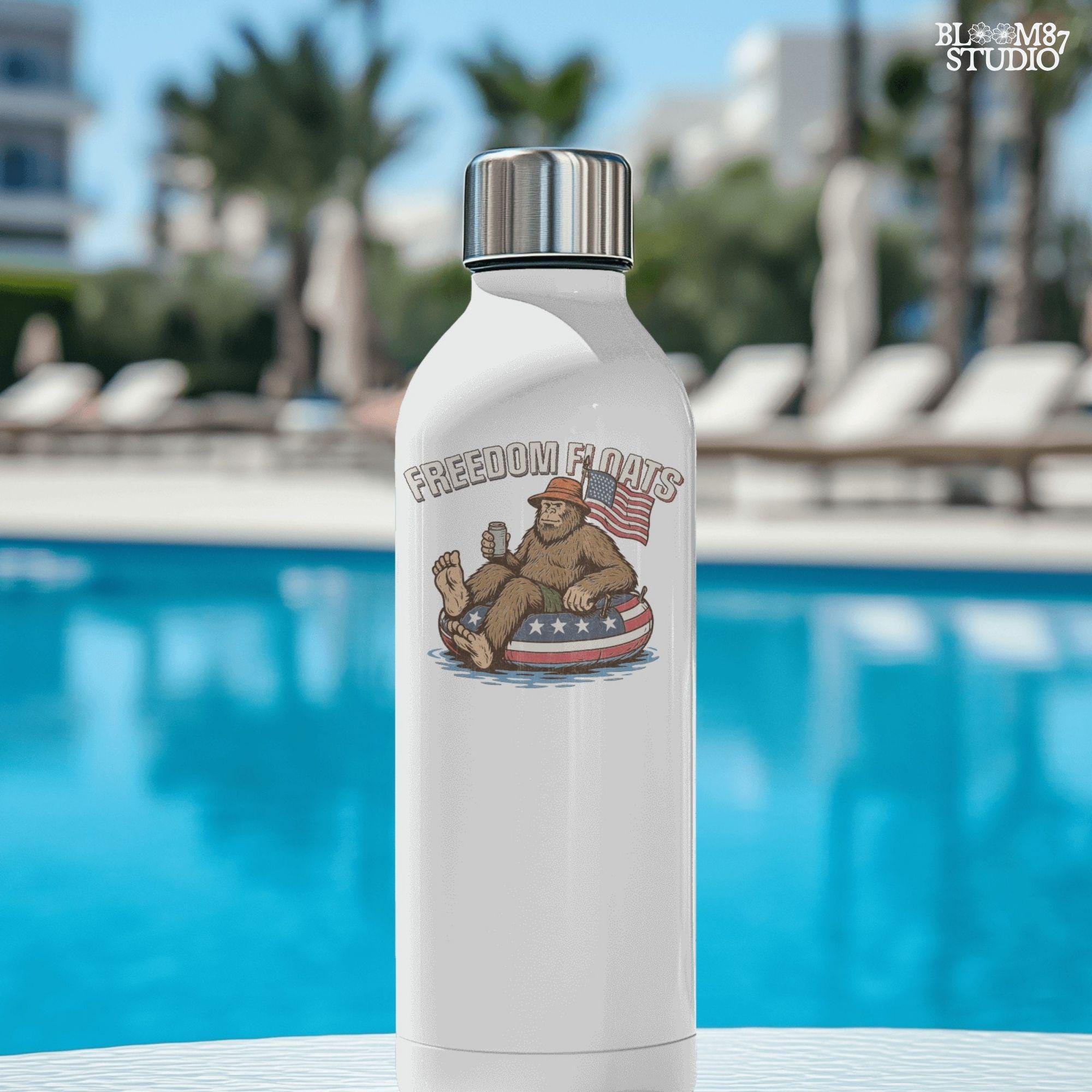 Bigfoot sitting on a patriotic float tube in water, holding a drink, wearing a hat, with an American flag behind and the phrase “Freedom Floats” above.