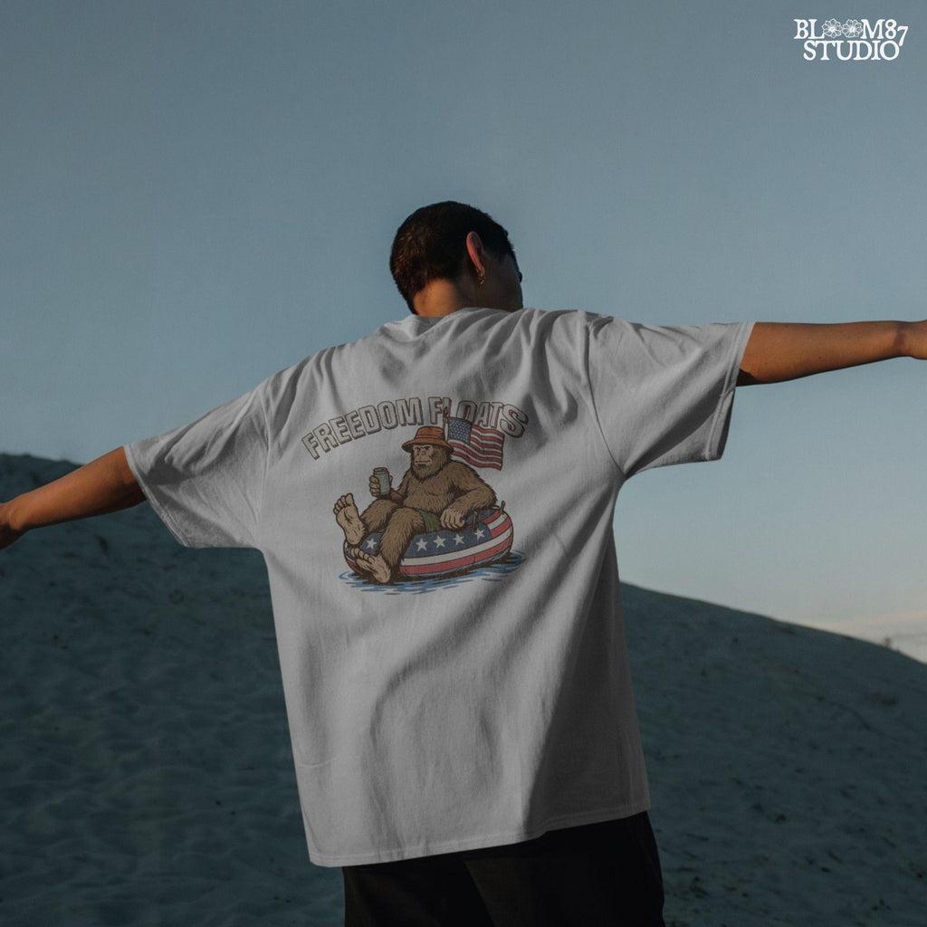Bigfoot sitting on a patriotic float tube in water, holding a drink, wearing a hat, with an American flag behind and the phrase “Freedom Floats” above.