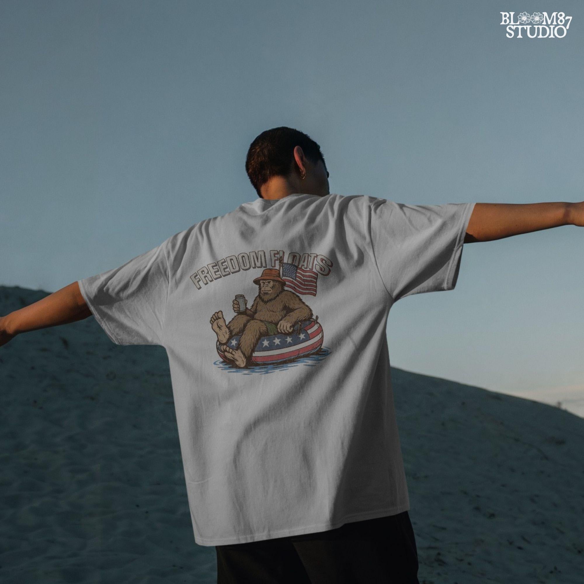 Bigfoot sitting on a patriotic float tube in water, holding a drink, wearing a hat, with an American flag behind and the phrase “Freedom Floats” above.