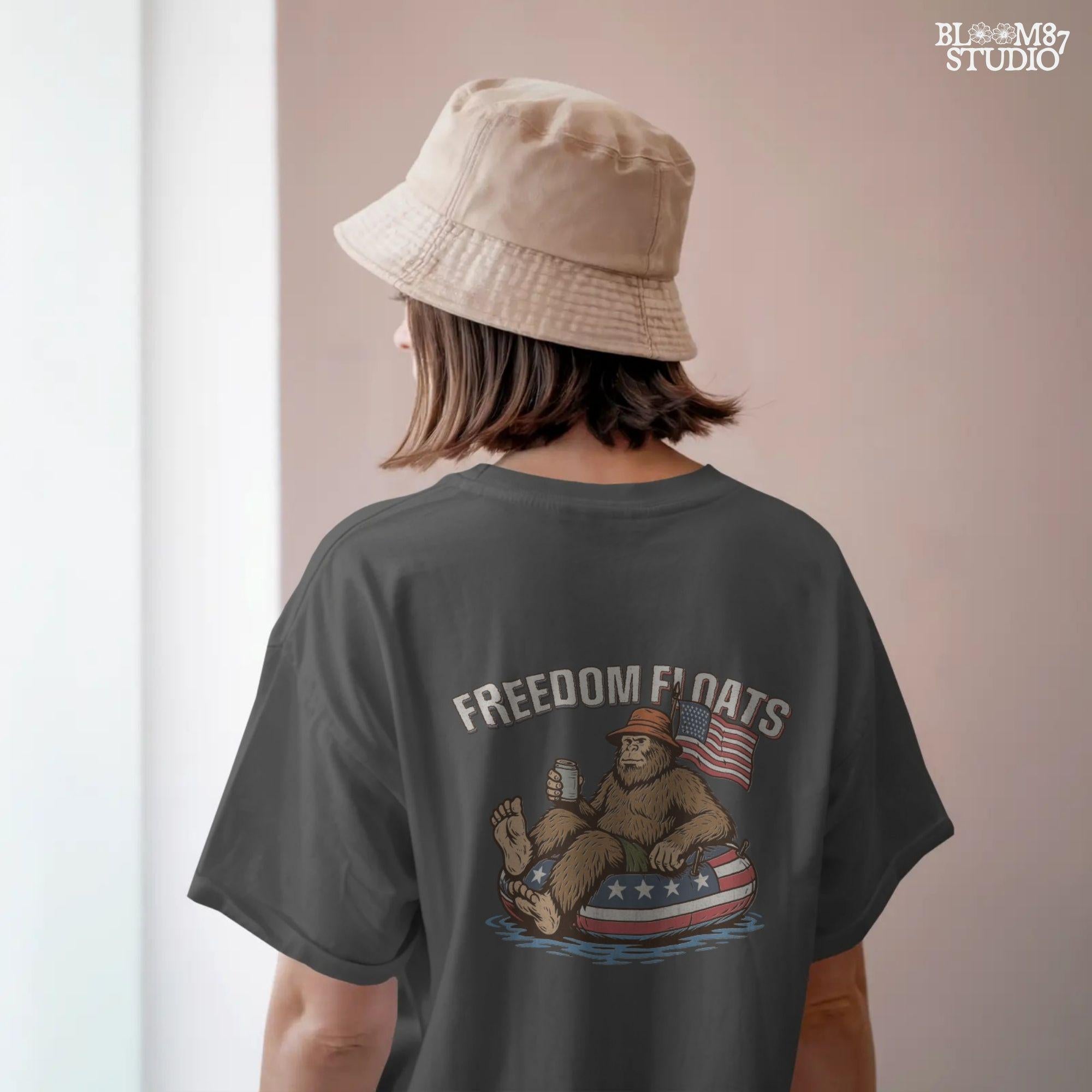Bigfoot sitting on a patriotic float tube in water, holding a drink, wearing a hat, with an American flag behind and the phrase “Freedom Floats” above.
