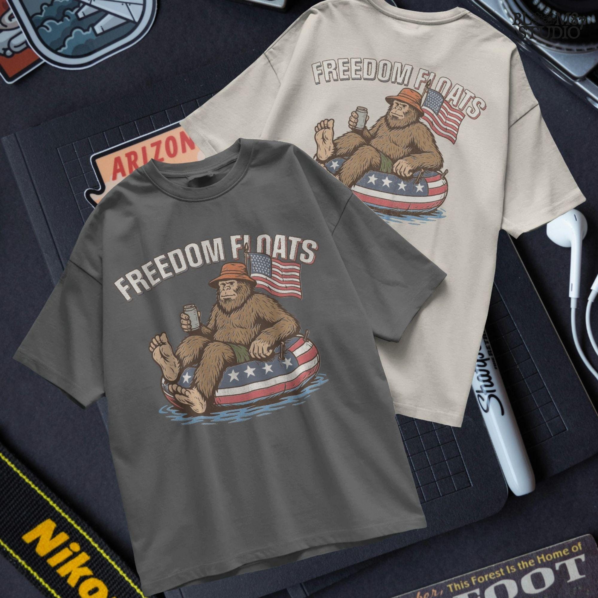 Bigfoot sitting on a patriotic float tube in water, holding a drink, wearing a hat, with an American flag behind and the phrase “Freedom Floats” above.