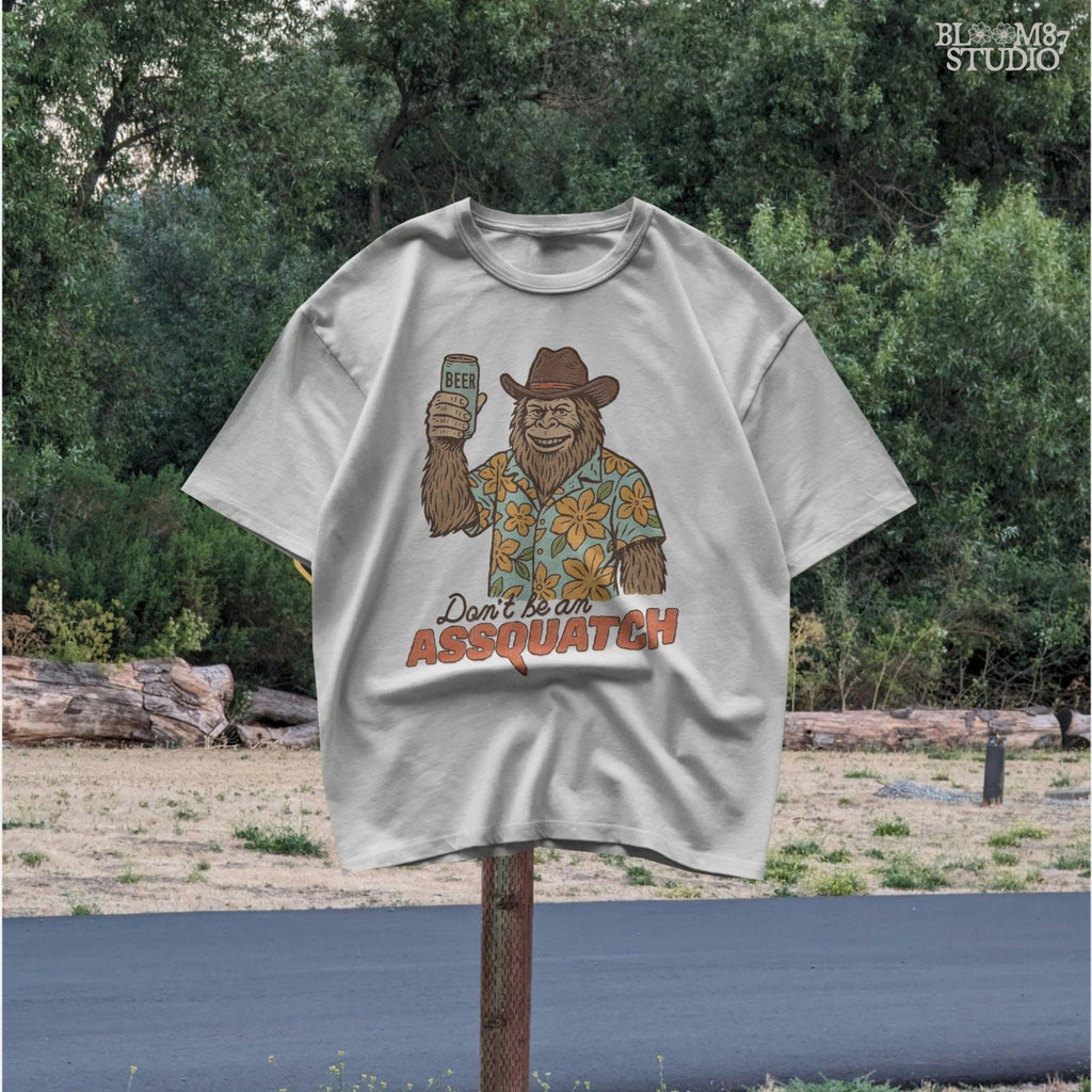 Retro cartoon Sasquatch in floral shirt holding a beer can with text “Don’t Be an Assquatch,” perfect for funny camping or summer designs