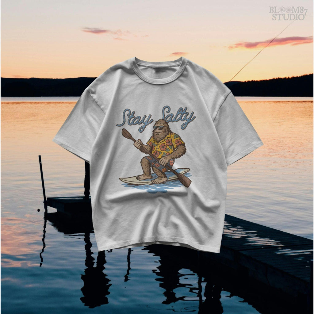 Cartoon Bigfoot wearing a Hawaiian shirt and sunglasses, paddleboarding on a surfboard with text “Stay Salty” – retro beach design PNG for sublimation.