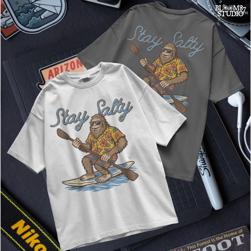 Cartoon Bigfoot wearing a Hawaiian shirt and sunglasses, paddleboarding on a surfboard with text “Stay Salty” – retro beach design PNG for sublimation.