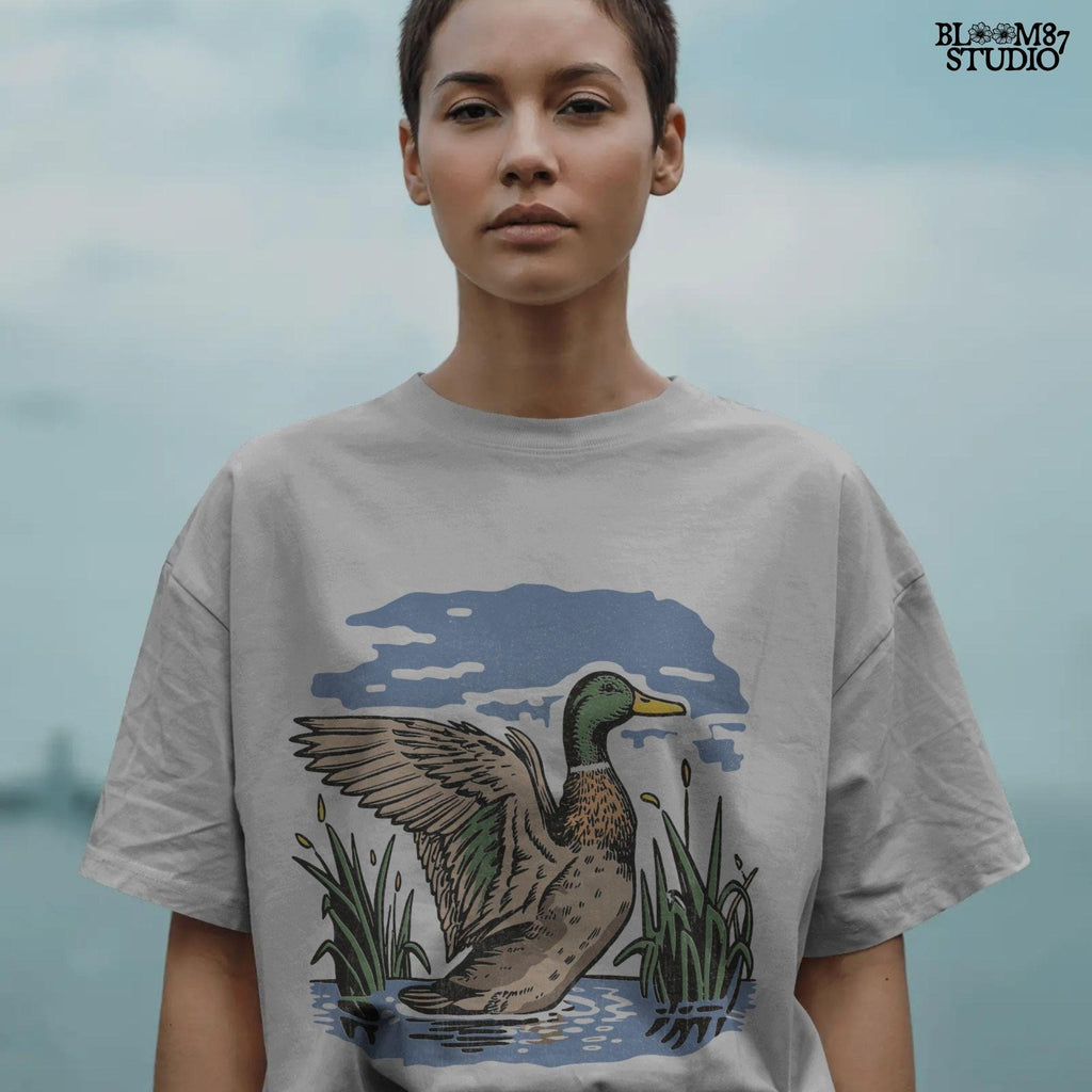 Vintage illustration of a mallard duck with wings spread standing in a pond with reeds and a cloudy blue sky – rustic nature sublimation PNG.