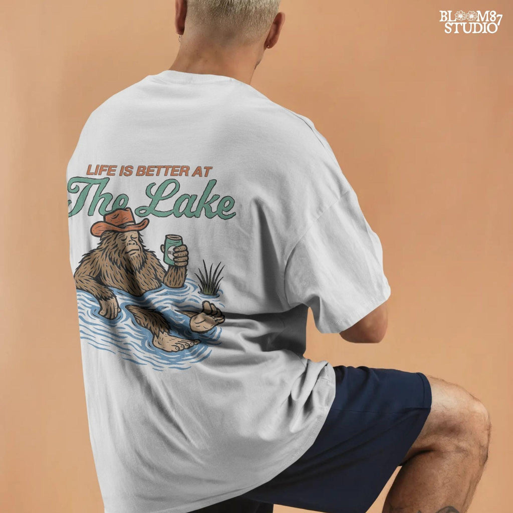 Cartoon Bigfoot relaxing in a lake wearing a cowboy hat, holding a beer can, with retro text saying “Life is Better at the Lake”