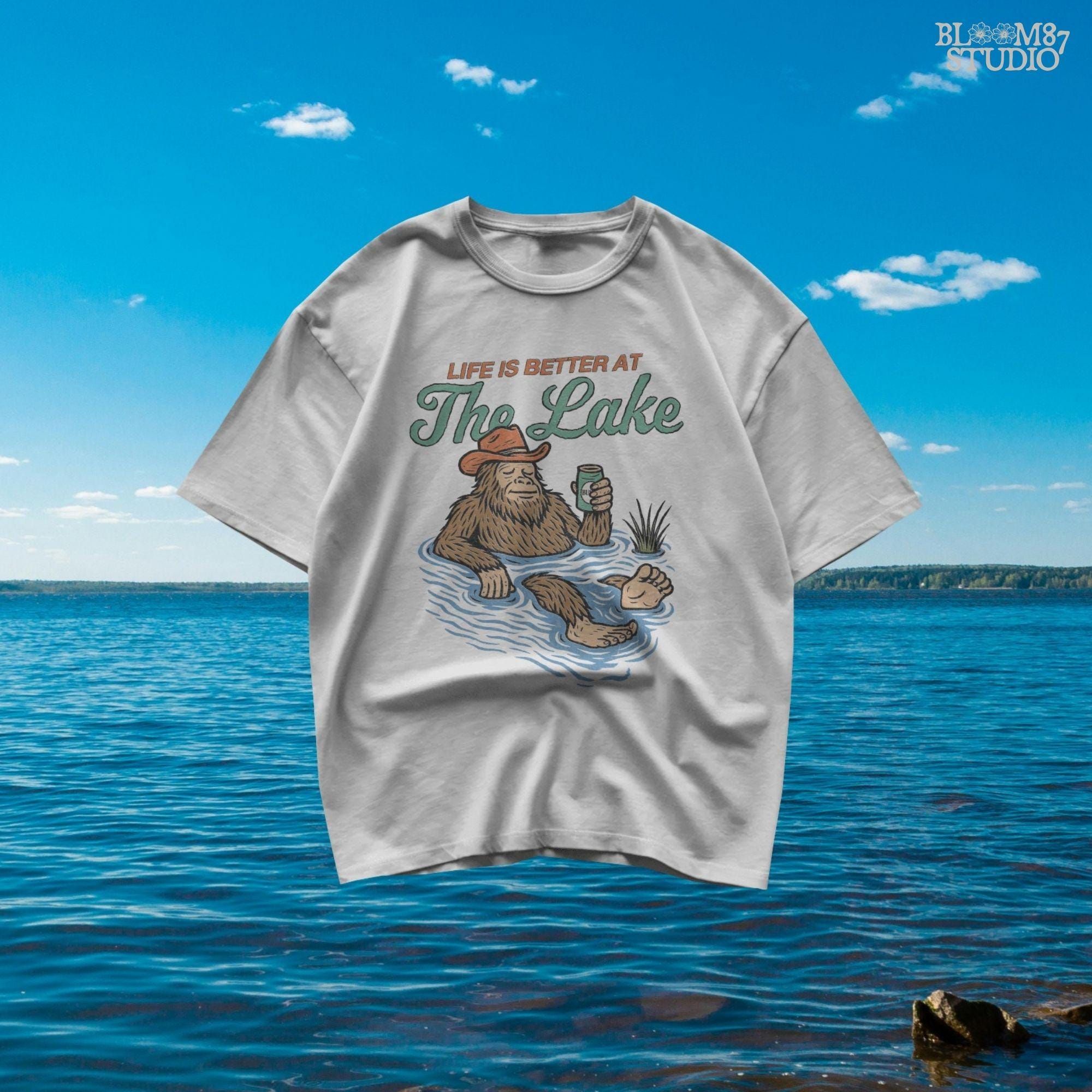 Cartoon Bigfoot relaxing in a lake wearing a cowboy hat, holding a beer can, with retro text saying “Life is Better at the Lake”