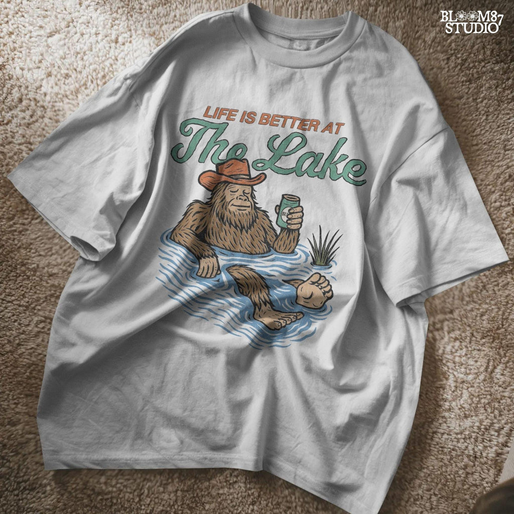 Cartoon Bigfoot relaxing in a lake wearing a cowboy hat, holding a beer can, with retro text saying “Life is Better at the Lake”