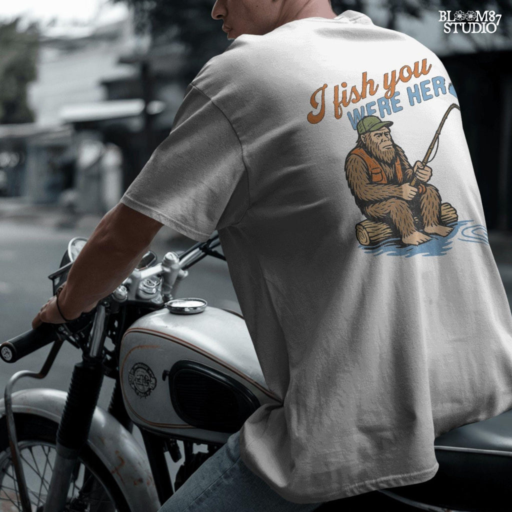 Funny Bigfoot character sitting on log fishing with bobber, wearing orange vest and green cap, with text “I Fish You Were Here”