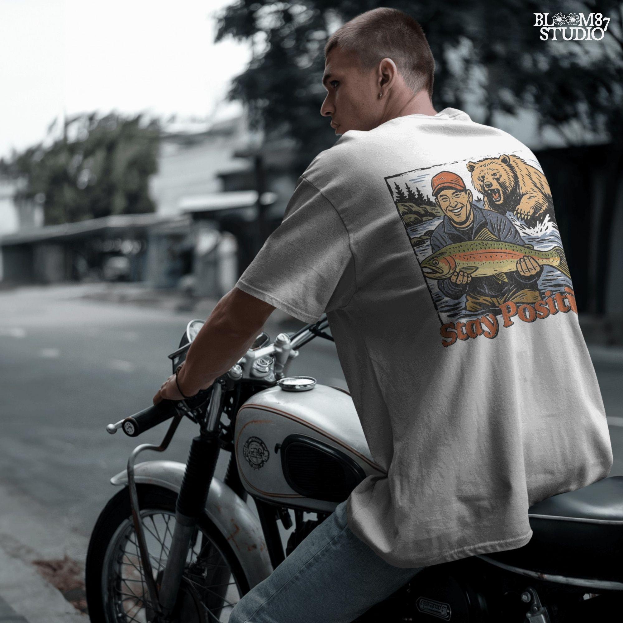 Happy fisherman holding large trout in river while a grizzly bear charges from behind, with text “Stay Positive” in funny outdoor design style