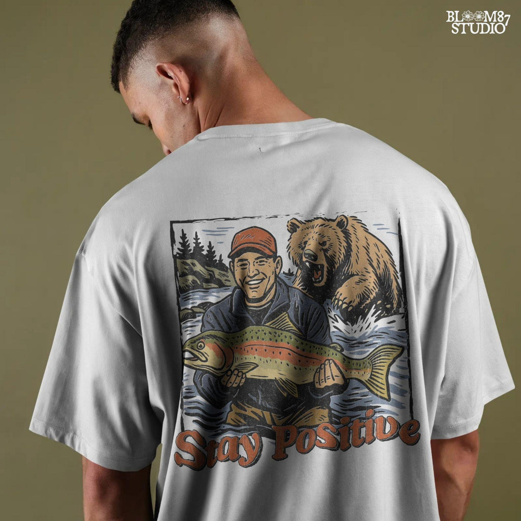 Happy fisherman holding large trout in river while a grizzly bear charges from behind, with text “Stay Positive” in funny outdoor design style