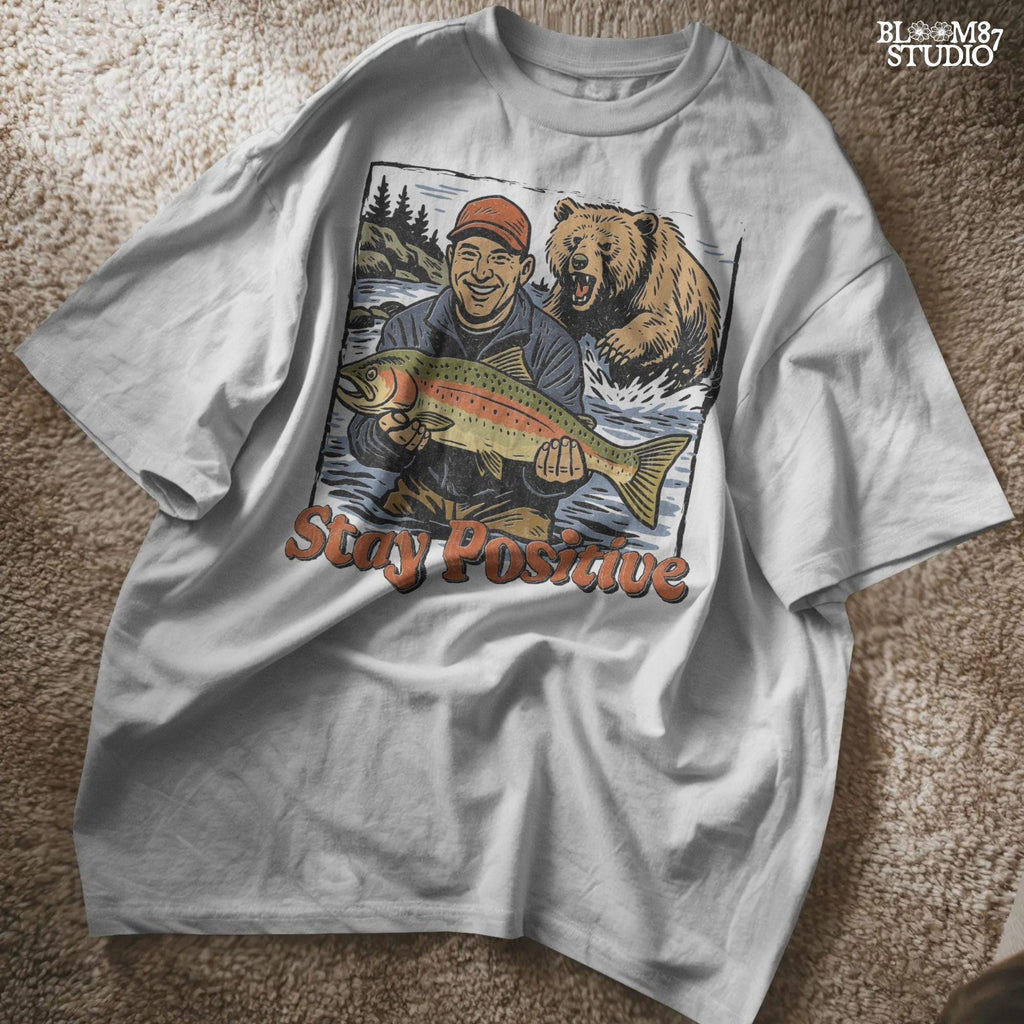 Happy fisherman holding large trout in river while a grizzly bear charges from behind, with text “Stay Positive” in funny outdoor design style