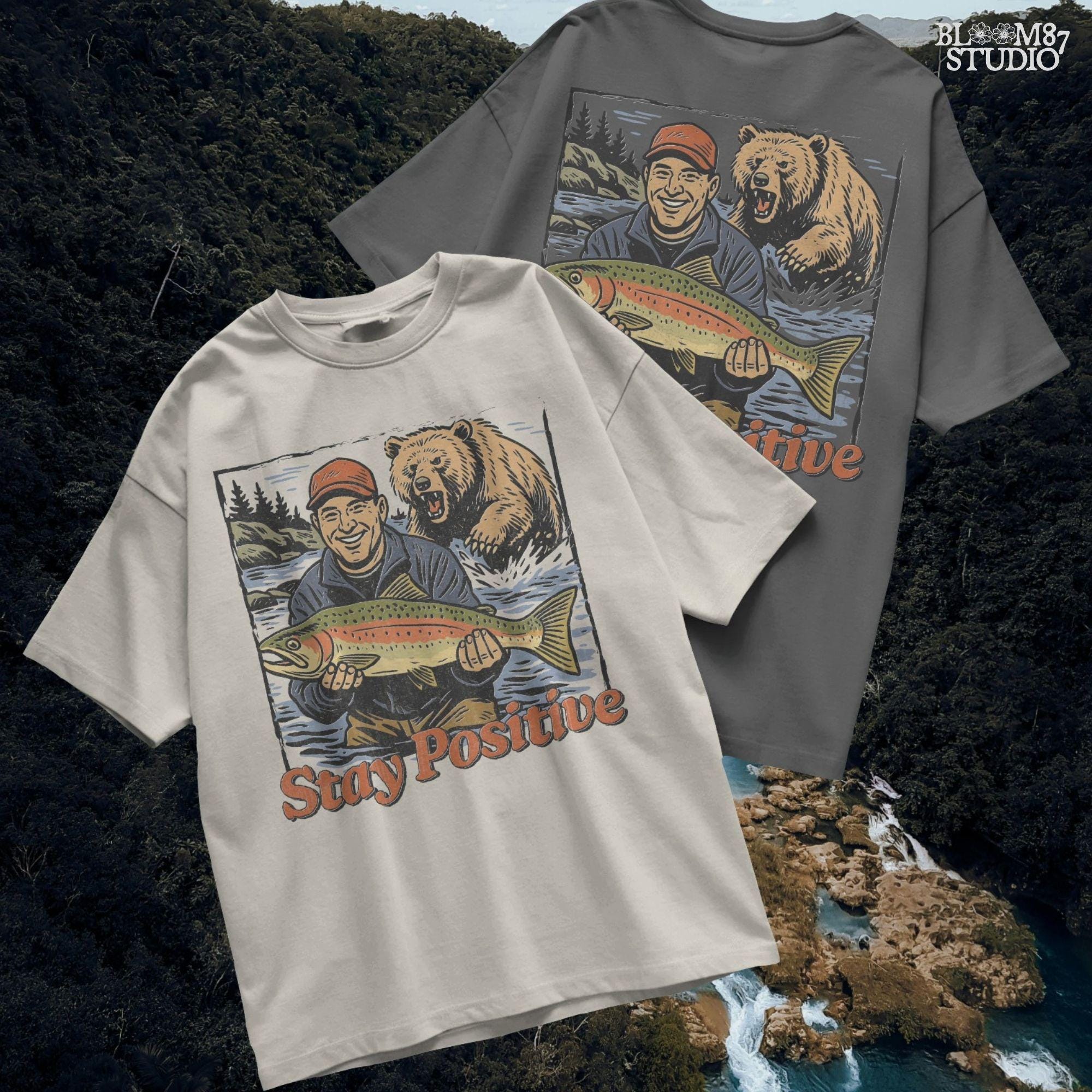 Happy fisherman holding large trout in river while a grizzly bear charges from behind, with text “Stay Positive” in funny outdoor design style