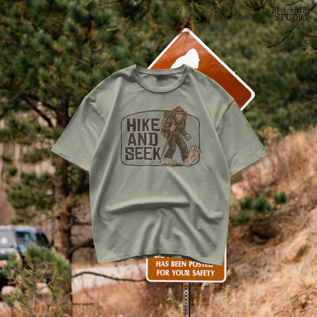 Illustration of a smiling Bigfoot wearing a ranger hat and hiking backpack with the phrase “Hike and Seek,” designed as a PNG for sublimation or print-on-demand products.