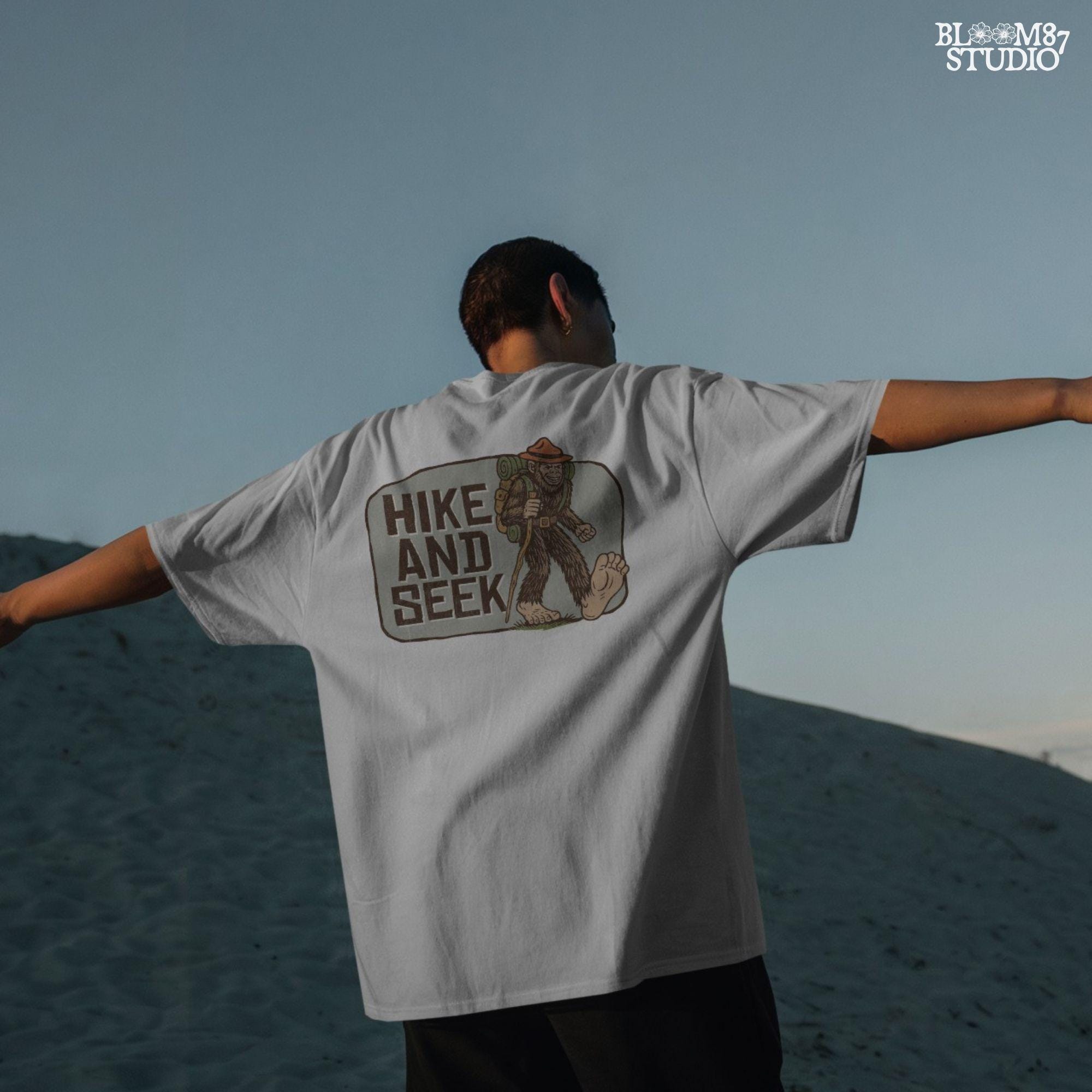 Illustration of a smiling Bigfoot wearing a ranger hat and hiking backpack with the phrase “Hike and Seek,” designed as a PNG for sublimation or print-on-demand products.