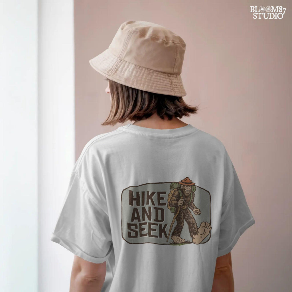 Illustration of a smiling Bigfoot wearing a ranger hat and hiking backpack with the phrase “Hike and Seek,” designed as a PNG for sublimation or print-on-demand products.