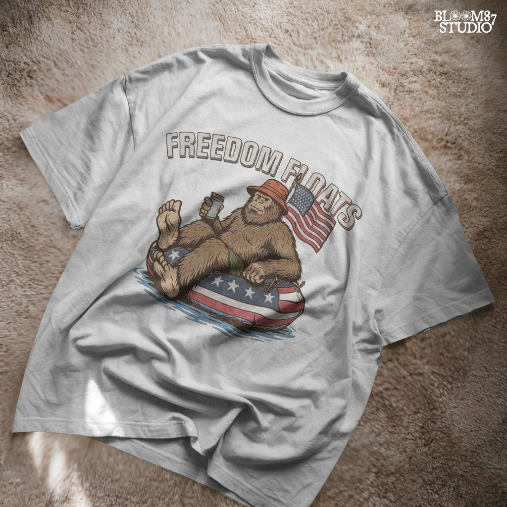 Bigfoot sitting on a patriotic float tube in water, holding a drink, wearing a hat, with an American flag behind and the phrase “Freedom Floats” above.