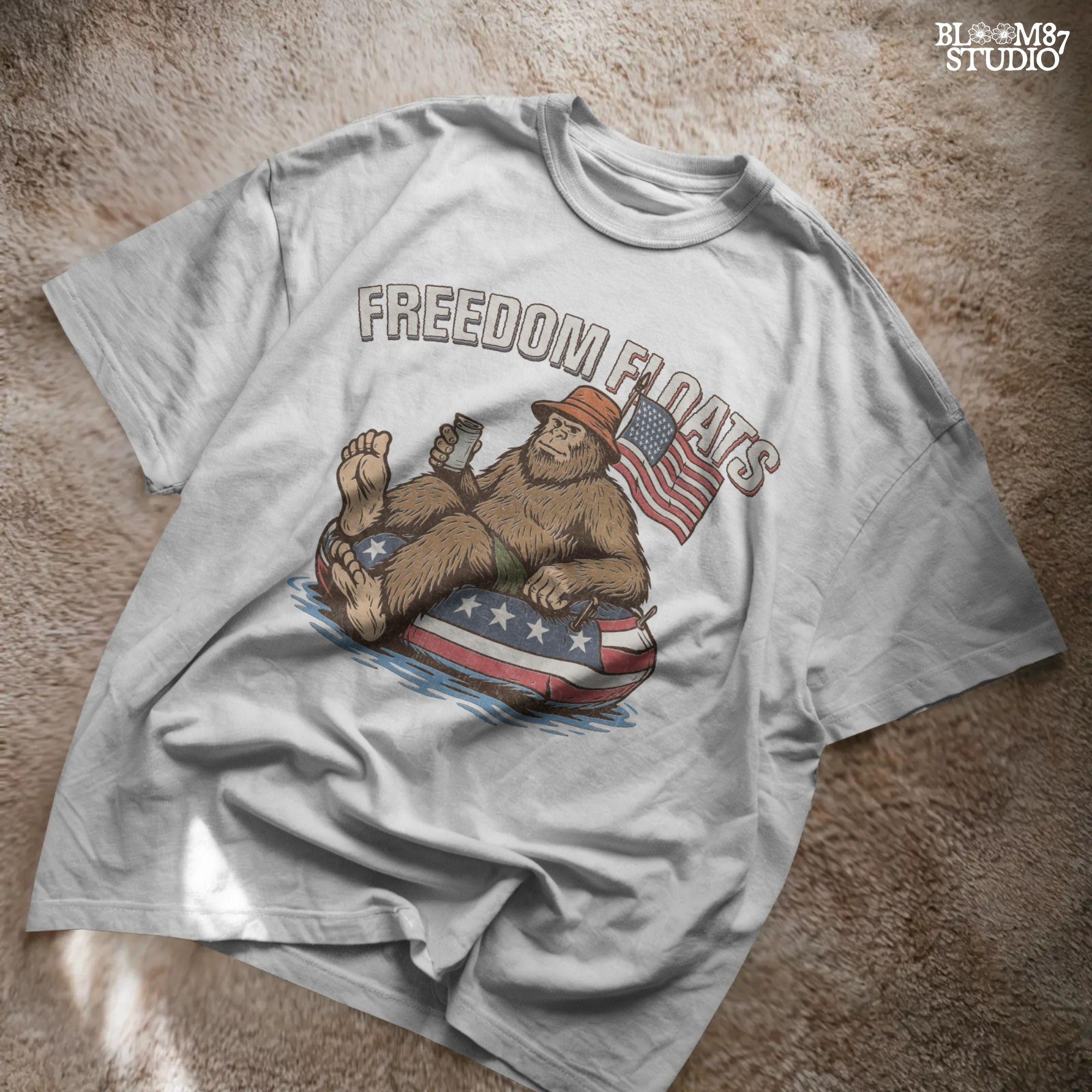 Bigfoot sitting on a patriotic float tube in water, holding a drink, wearing a hat, with an American flag behind and the phrase “Freedom Floats” above.