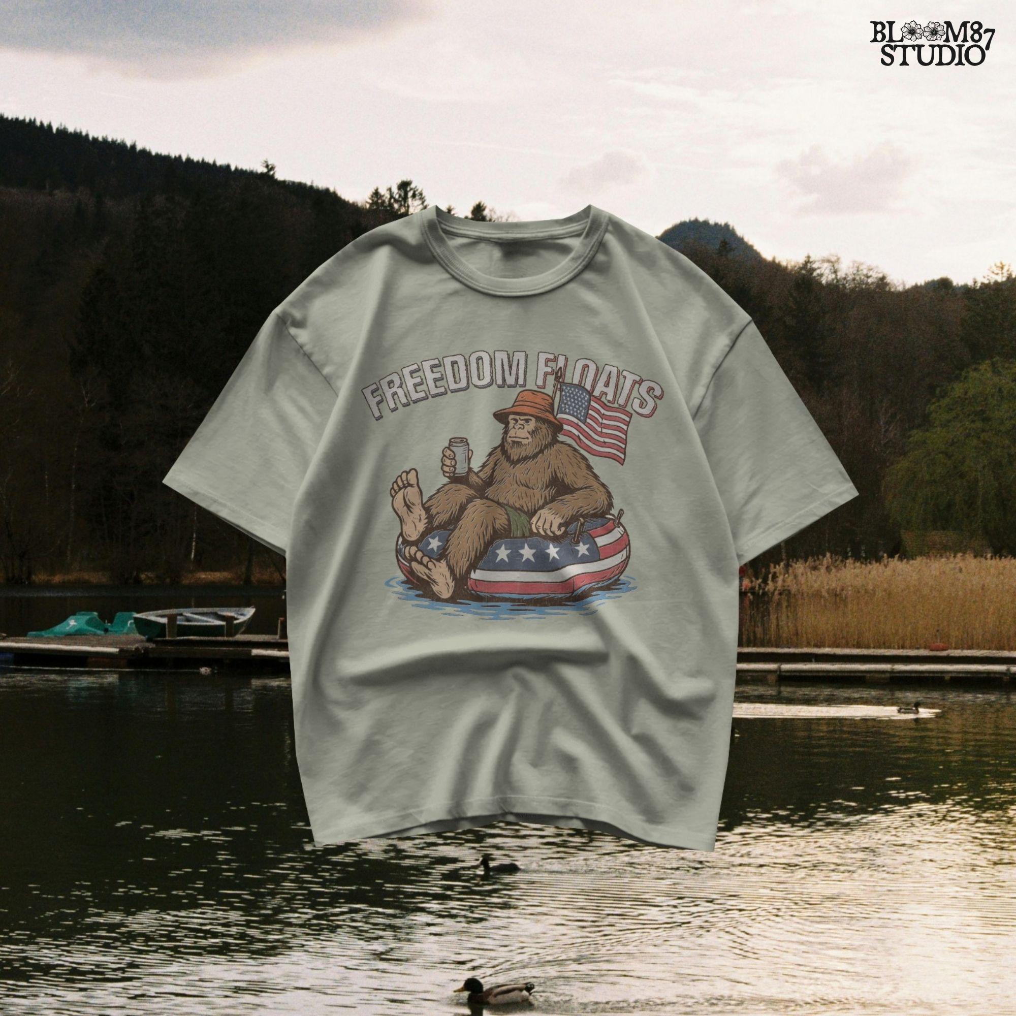 Bigfoot sitting on a patriotic float tube in water, holding a drink, wearing a hat, with an American flag behind and the phrase “Freedom Floats” above.