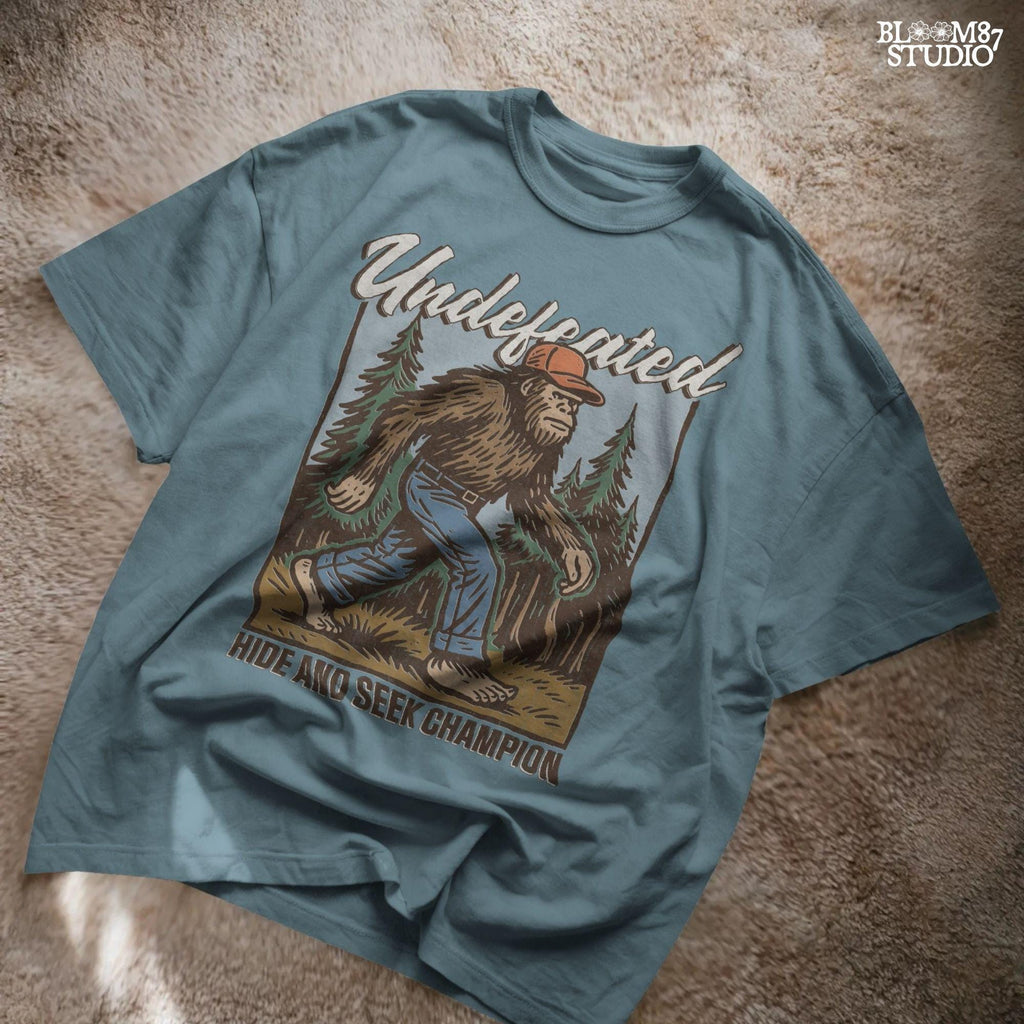 Hand-drawn Bigfoot walking through a pine forest in a red cap and jeans, with text “Undefeated Hide and Seek Champion,” designed for sublimation use.