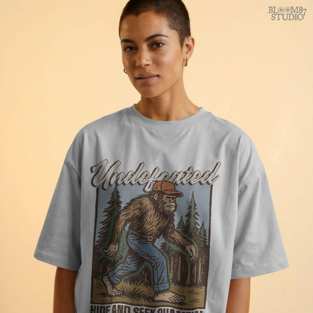 Hand-drawn Bigfoot walking through a pine forest in a red cap and jeans, with text “Undefeated Hide and Seek Champion,” designed for sublimation use.