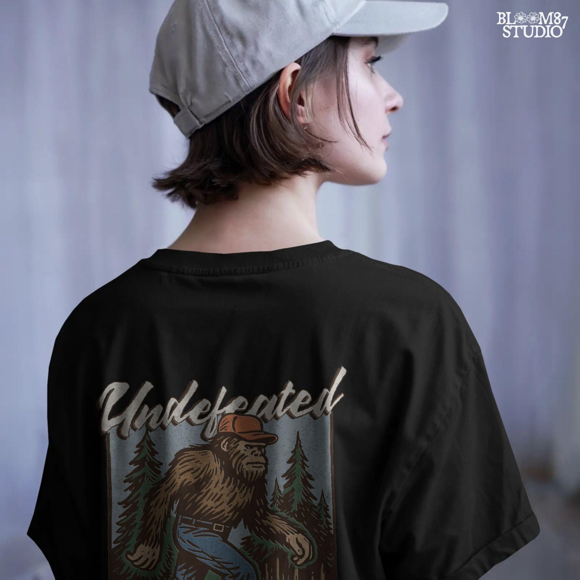 Hand-drawn Bigfoot walking through a pine forest in a red cap and jeans, with text “Undefeated Hide and Seek Champion,” designed for sublimation use.