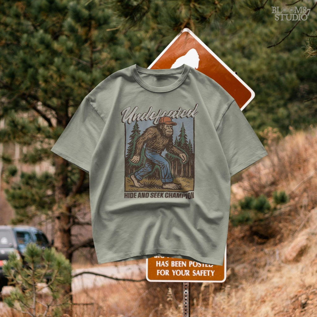 Hand-drawn Bigfoot walking through a pine forest in a red cap and jeans, with text “Undefeated Hide and Seek Champion,” designed for sublimation use.