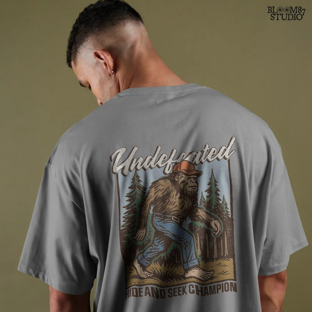 Hand-drawn Bigfoot walking through a pine forest in a red cap and jeans, with text “Undefeated Hide and Seek Champion,” designed for sublimation use.
