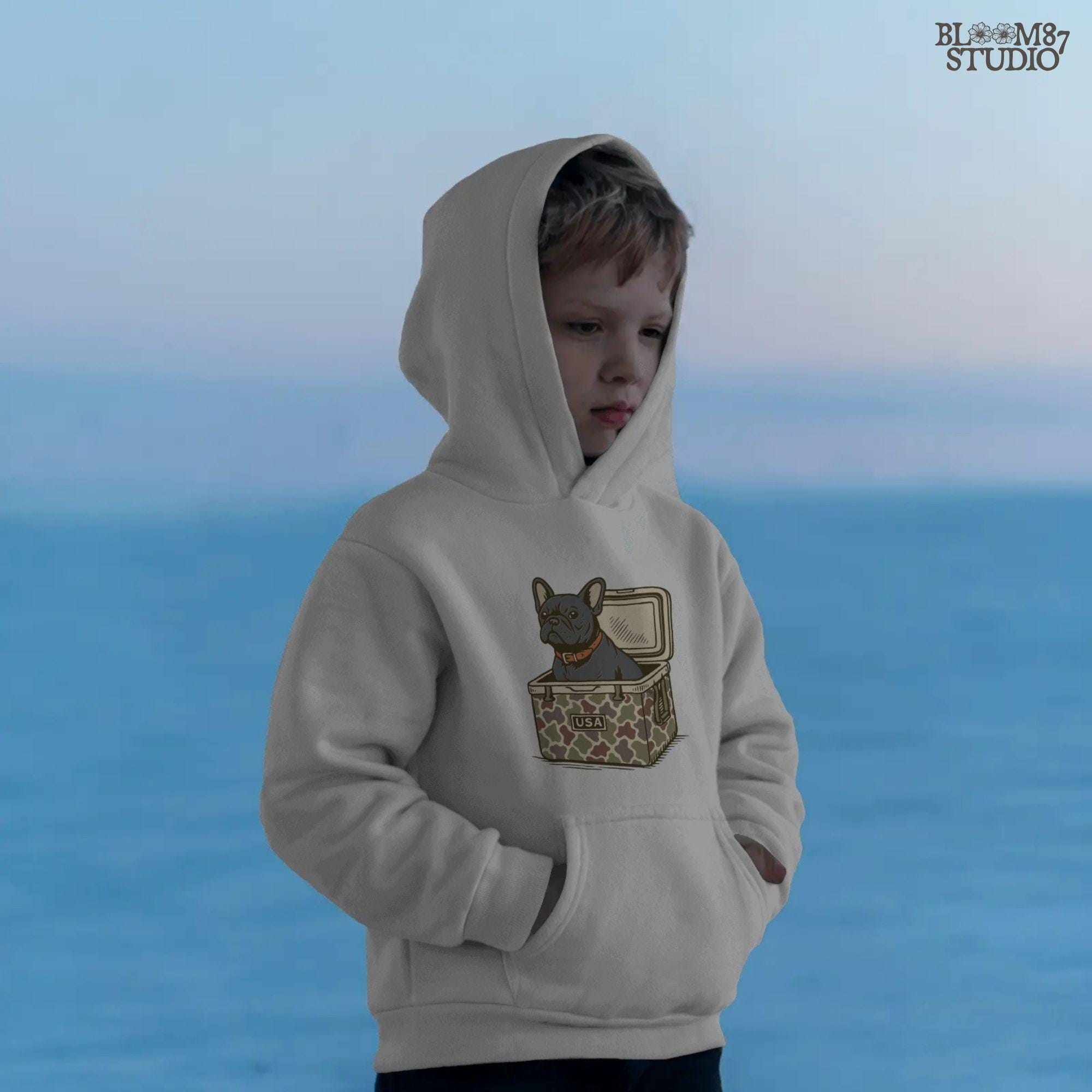 French Bulldog sitting inside a camouflage USA cooler, wearing a red collar, in rustic outdoor style — perfect for patriotic dog lovers and sublimation use.