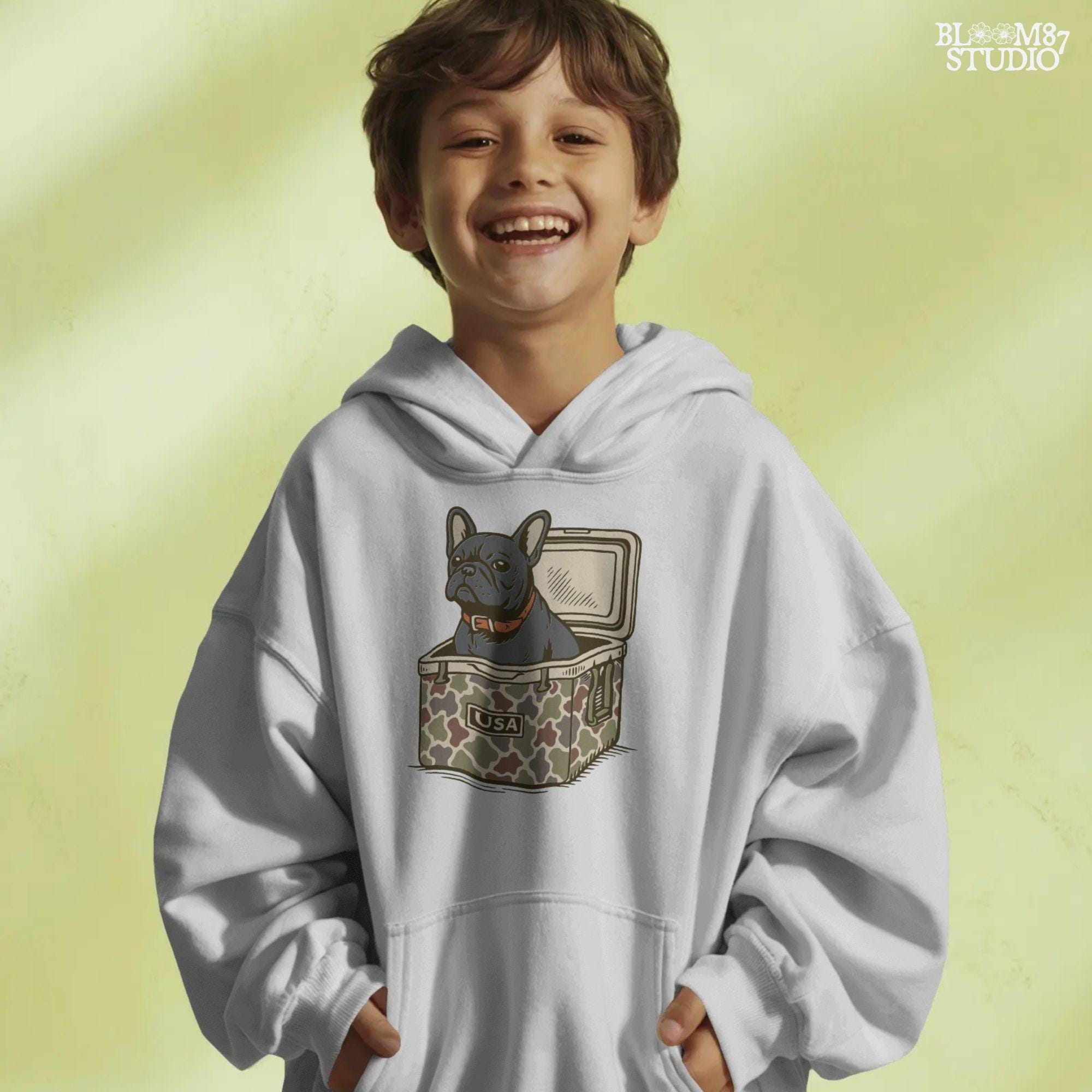 French Bulldog sitting inside a camouflage USA cooler, wearing a red collar, in rustic outdoor style — perfect for patriotic dog lovers and sublimation use.