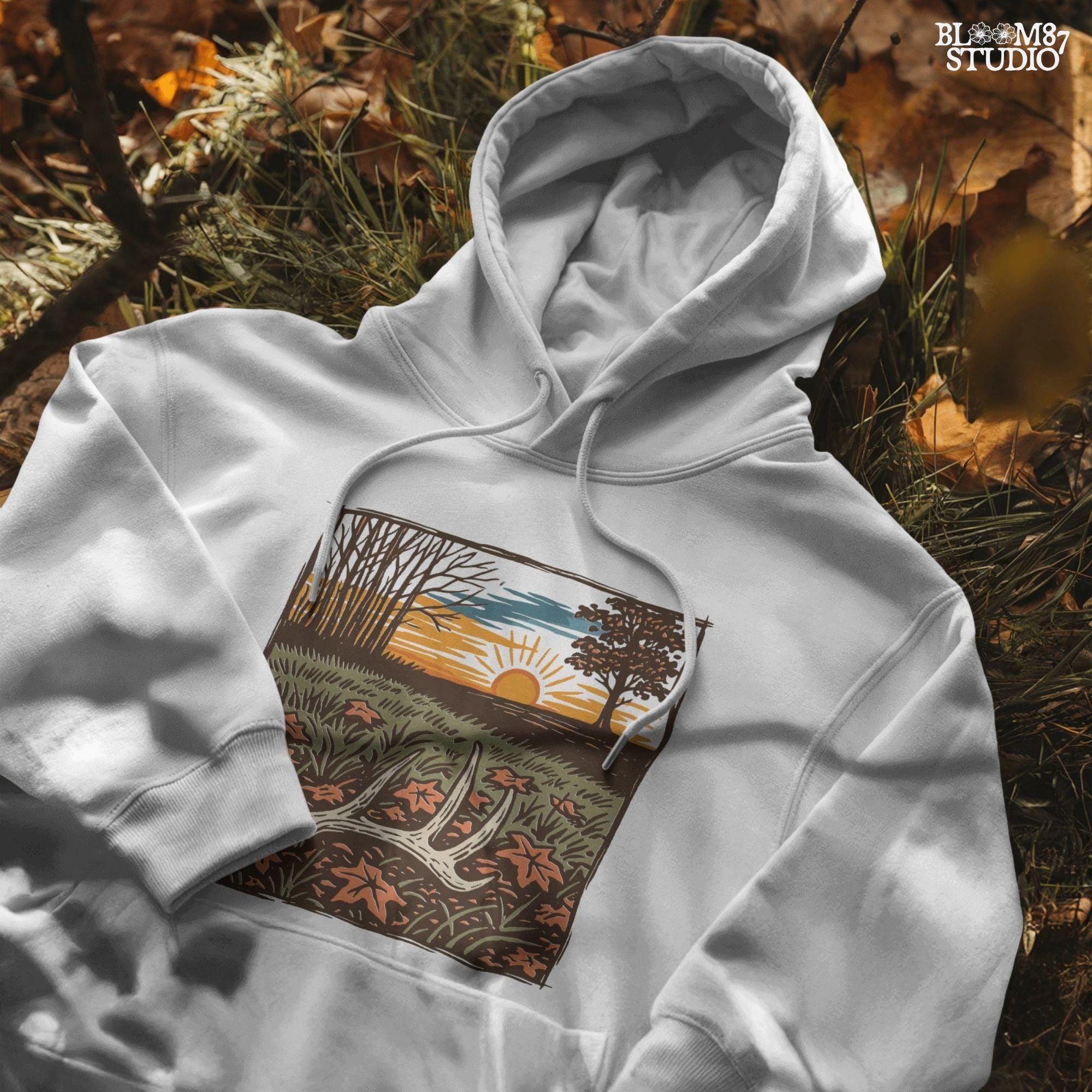 A rustic illustration of a whitetail antler lying in a fall field with orange leaves and a vibrant sunset sky in the background, PNG format for sublimation.