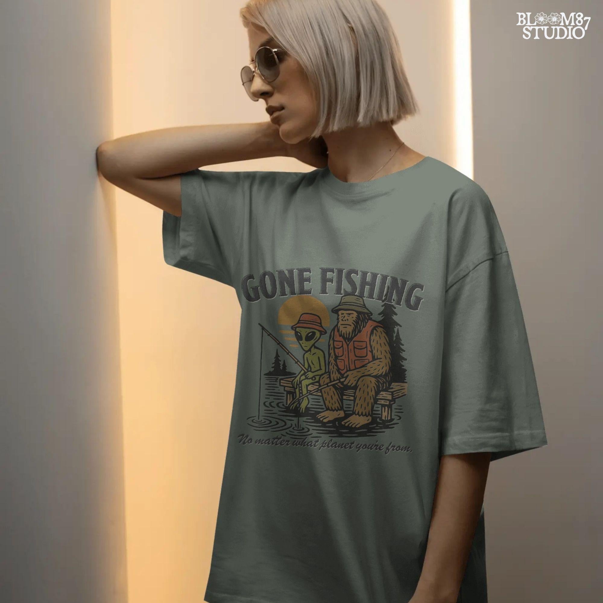 Funny illustration of Bigfoot and an alien fishing together on a lake dock at sunset, wearing fishing hats and vests—perfect for summer and Father’s Day sublimation.