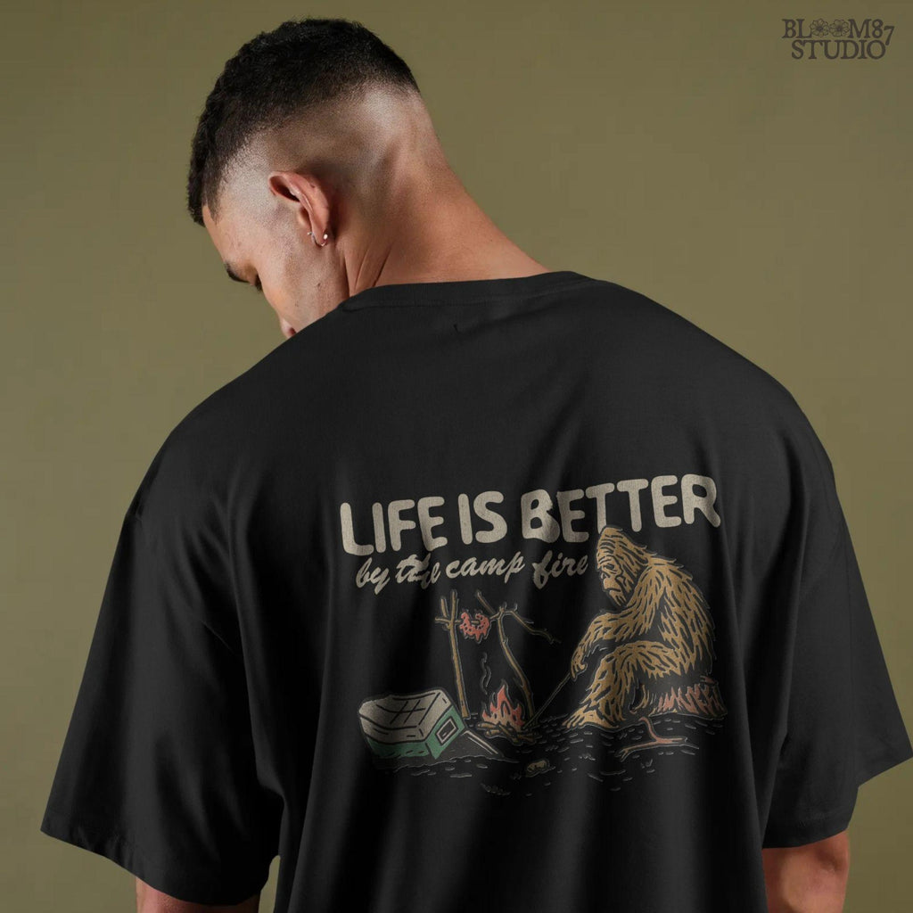 Bigfoot roasting meat over a campfire next to a tipped cooler, with the phrase “Life Is Better by the camp fire” above, illustrated PNG design for outdoor sublimation.