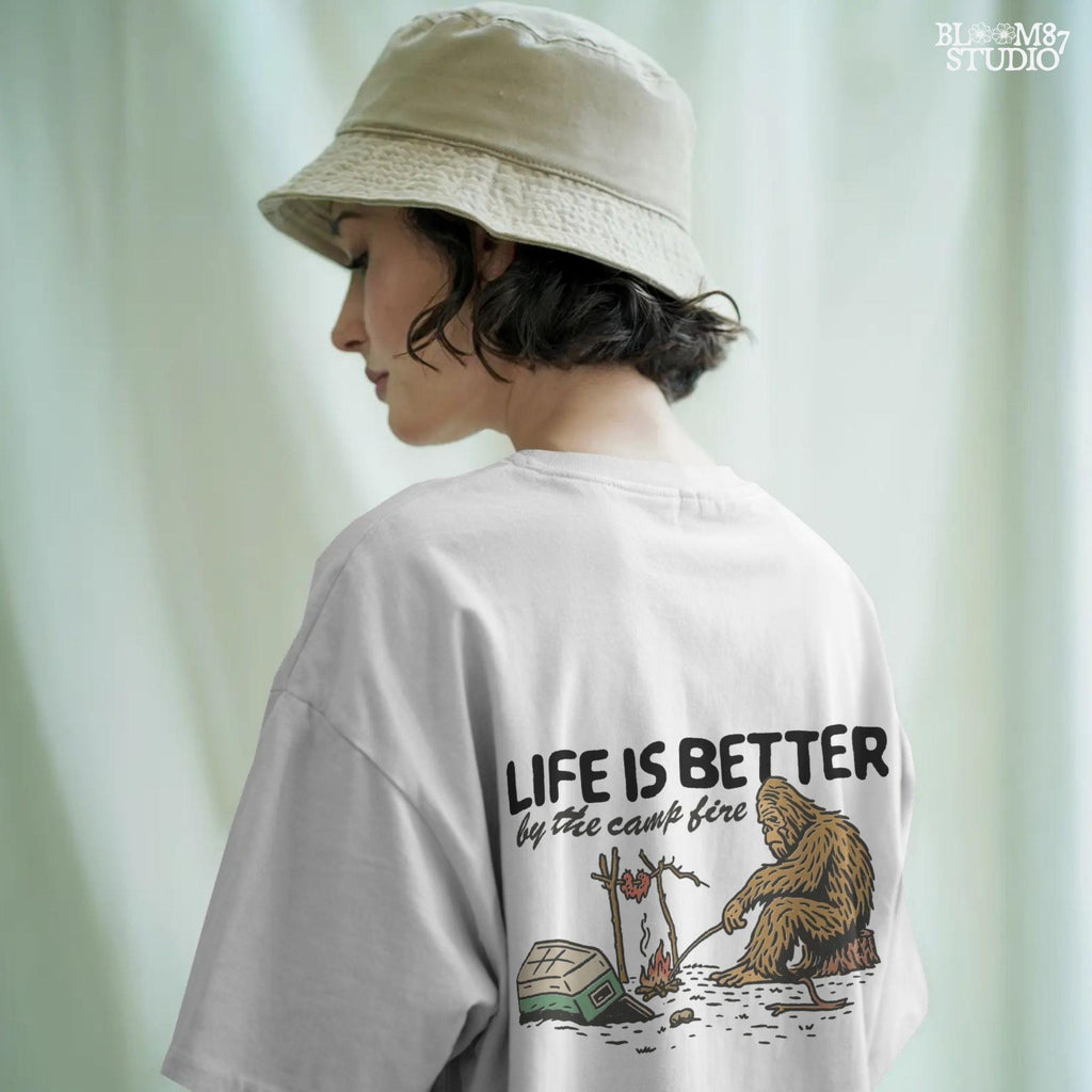 Bigfoot roasting meat over a campfire next to a tipped cooler, with the phrase “Life Is Better by the camp fire” above, illustrated PNG design for outdoor sublimation.